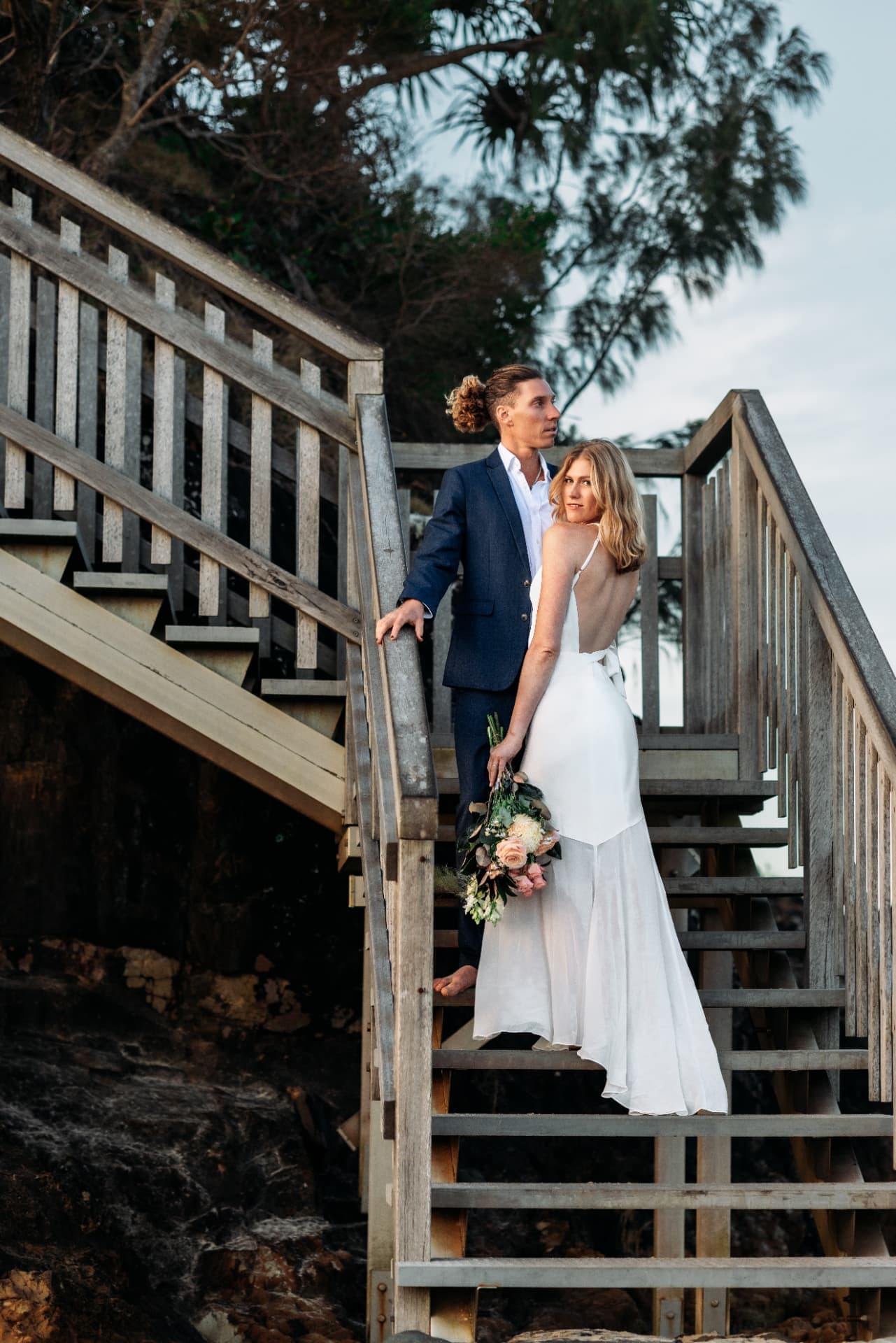 Chloe and Jojo standing together on weathered cliffside timber stairs, Chloe glancing back over her shoulder