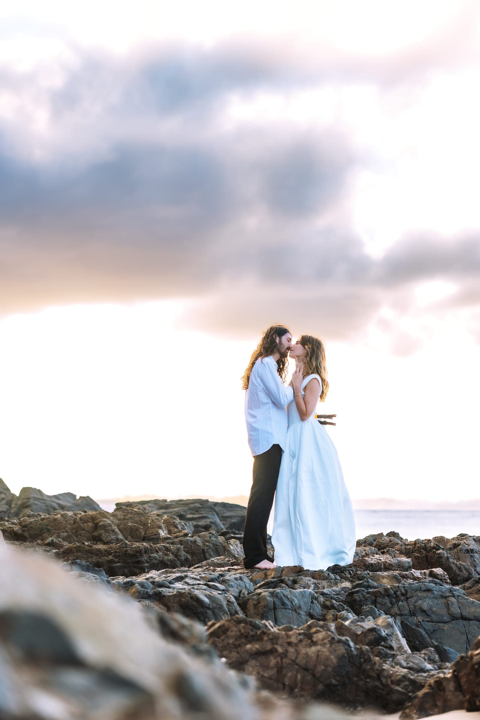Couple sharing a kiss on the coastal rocks beneath a soft cloudy sunrise