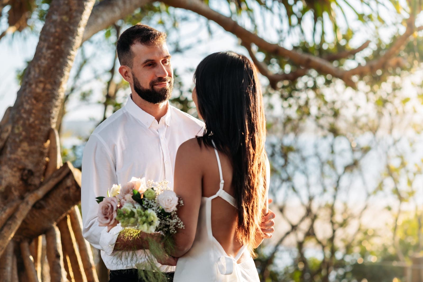 Groom smiling at his bride beneath a pandanus tree in golden light