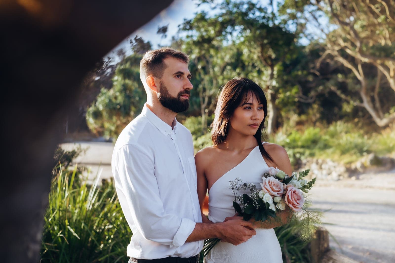 Couple standing together in the coastal bush, the bride holding a pink rose bouquet