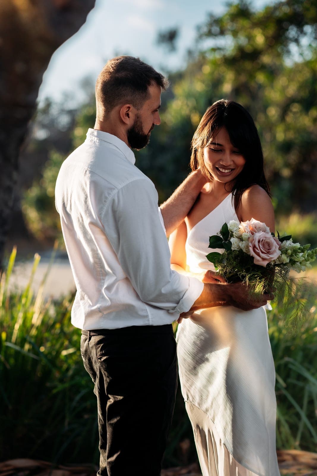 Bride smiling down at her bouquet as the groom touches her neck in golden backlight