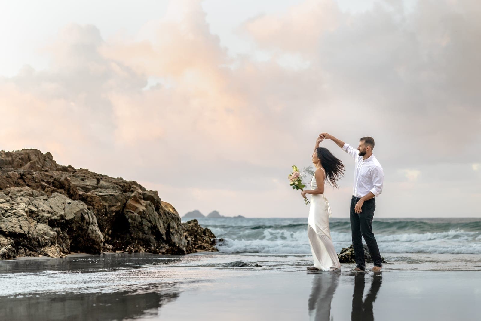 Groom twirling his bride on the wet sand beside the rocks under a pink sunset sky