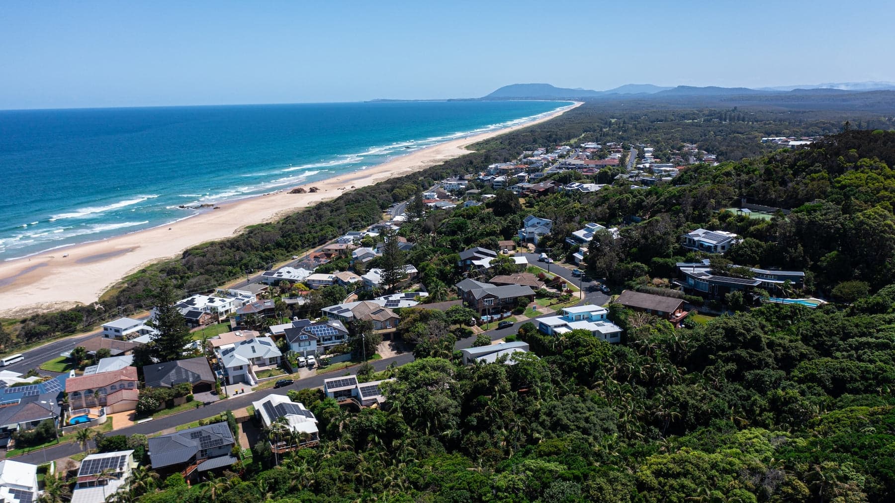 Aerial of a long surf beach with bushland homes and mountains in the distance