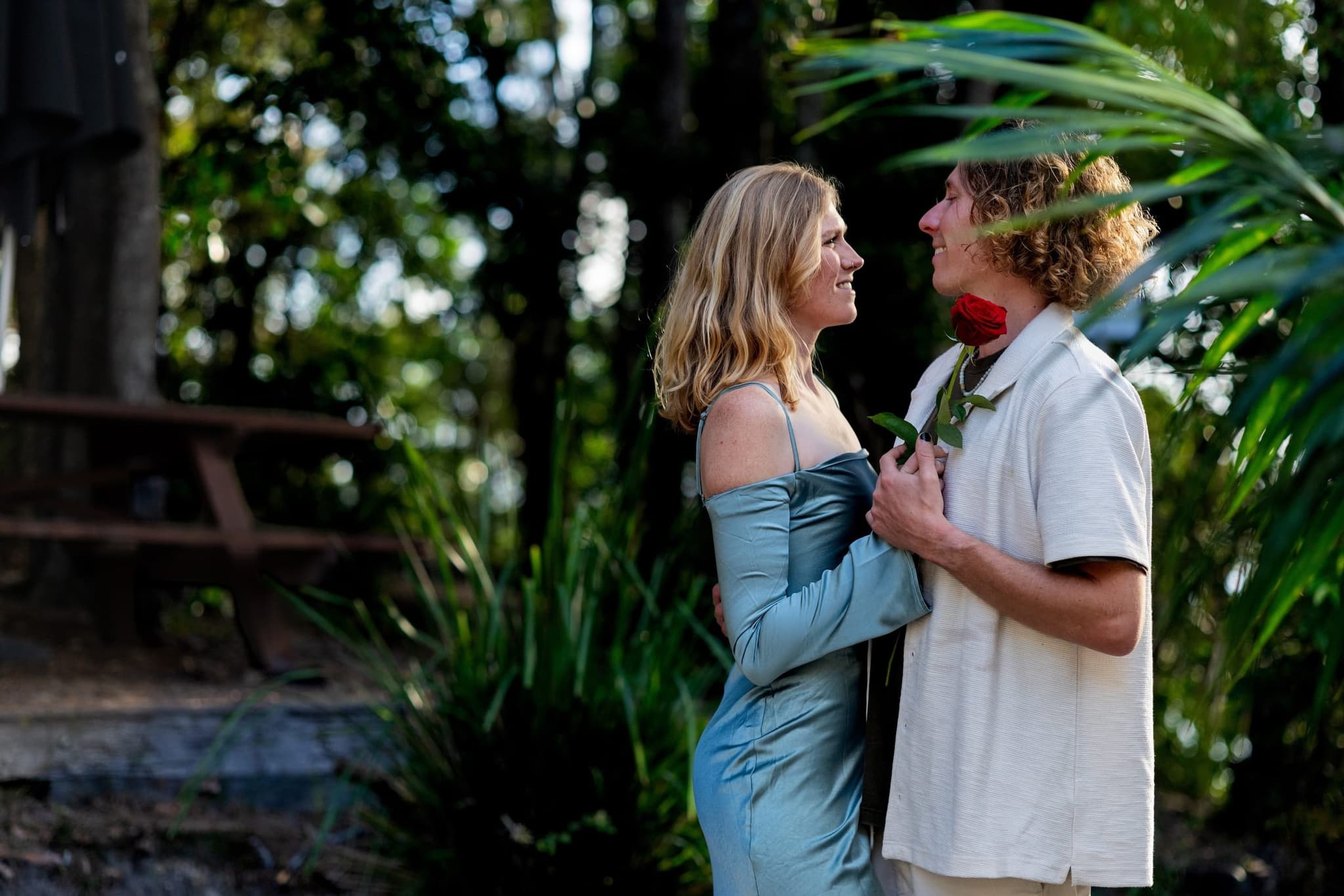 Chloe and JoJo smiling at each other among palm fronds, a red rose held between them