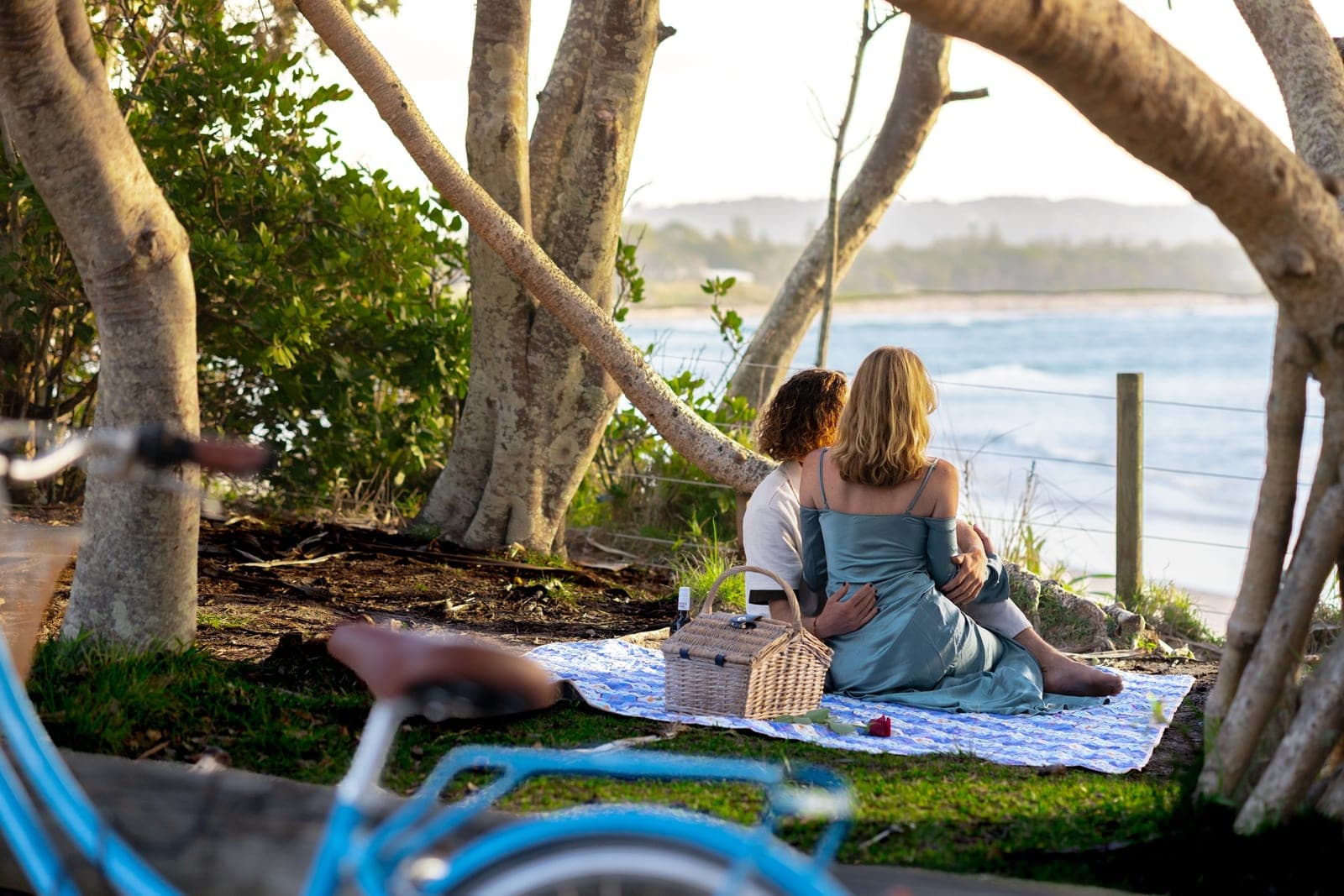 Chloe and JoJo on a picnic blanket overlooking the ocean, a blue bicycle in the foreground