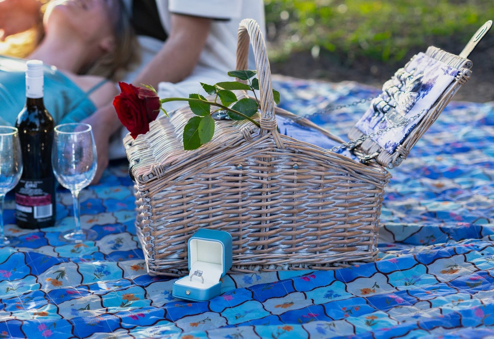 An open ring box beside a wicker picnic basket topped with a red rose