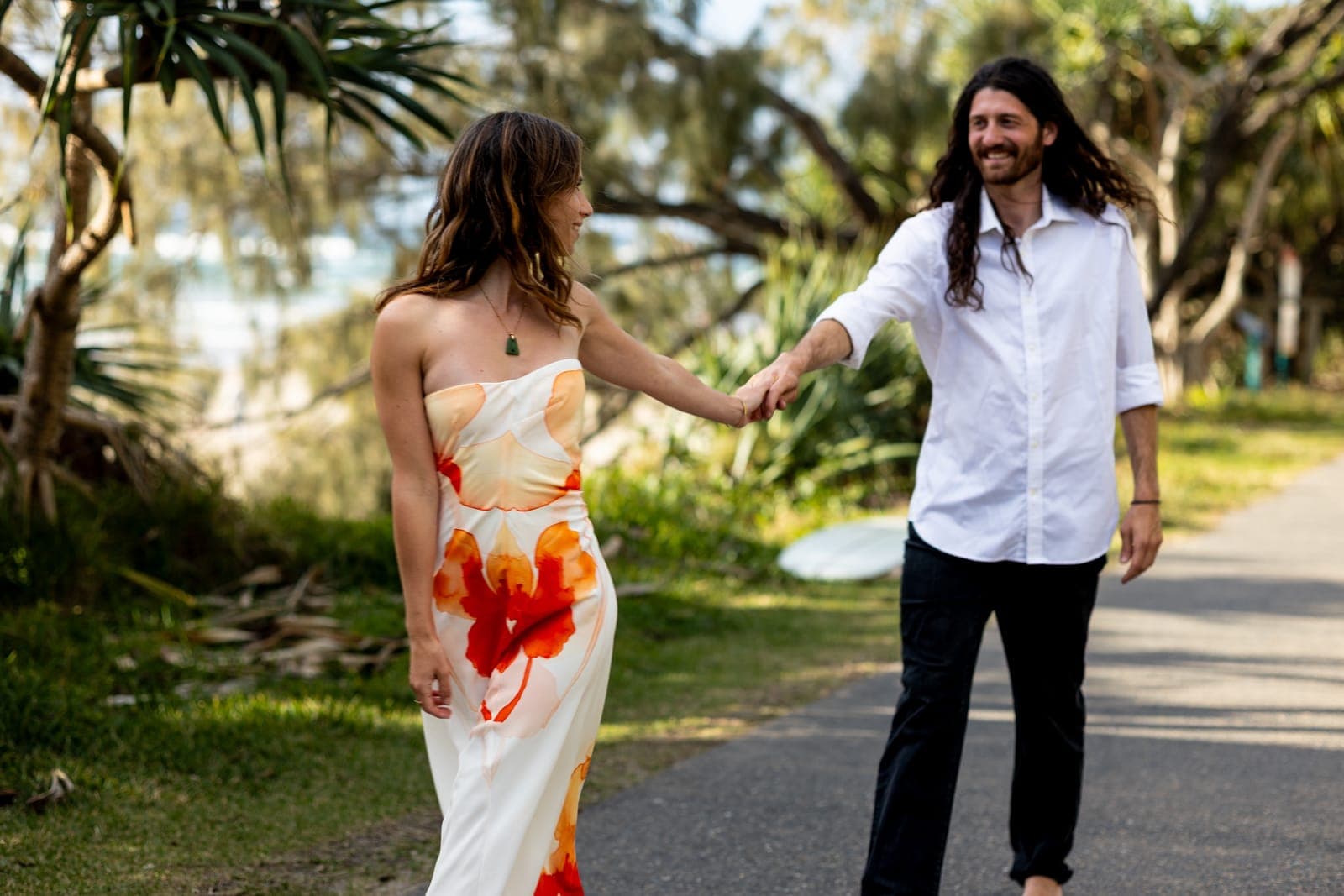 Charlene leading Jerome by the hand along a coastal path under pandanus trees