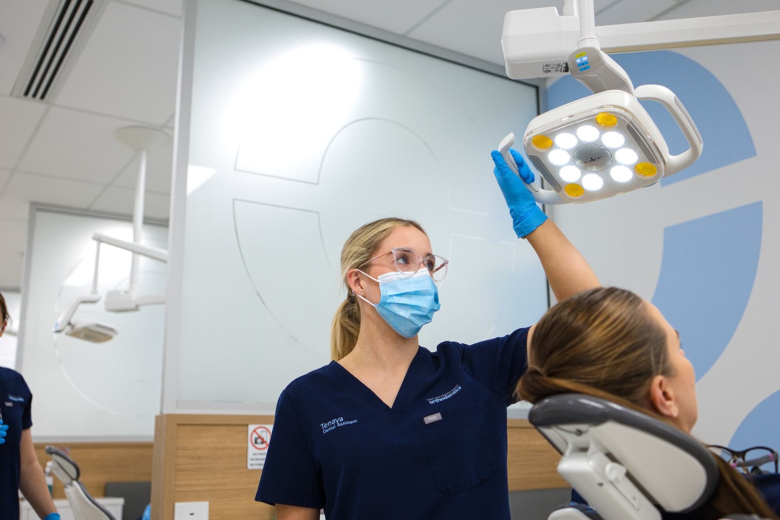 Dental assistant in navy scrubs and mask adjusting the overhead light above a patient