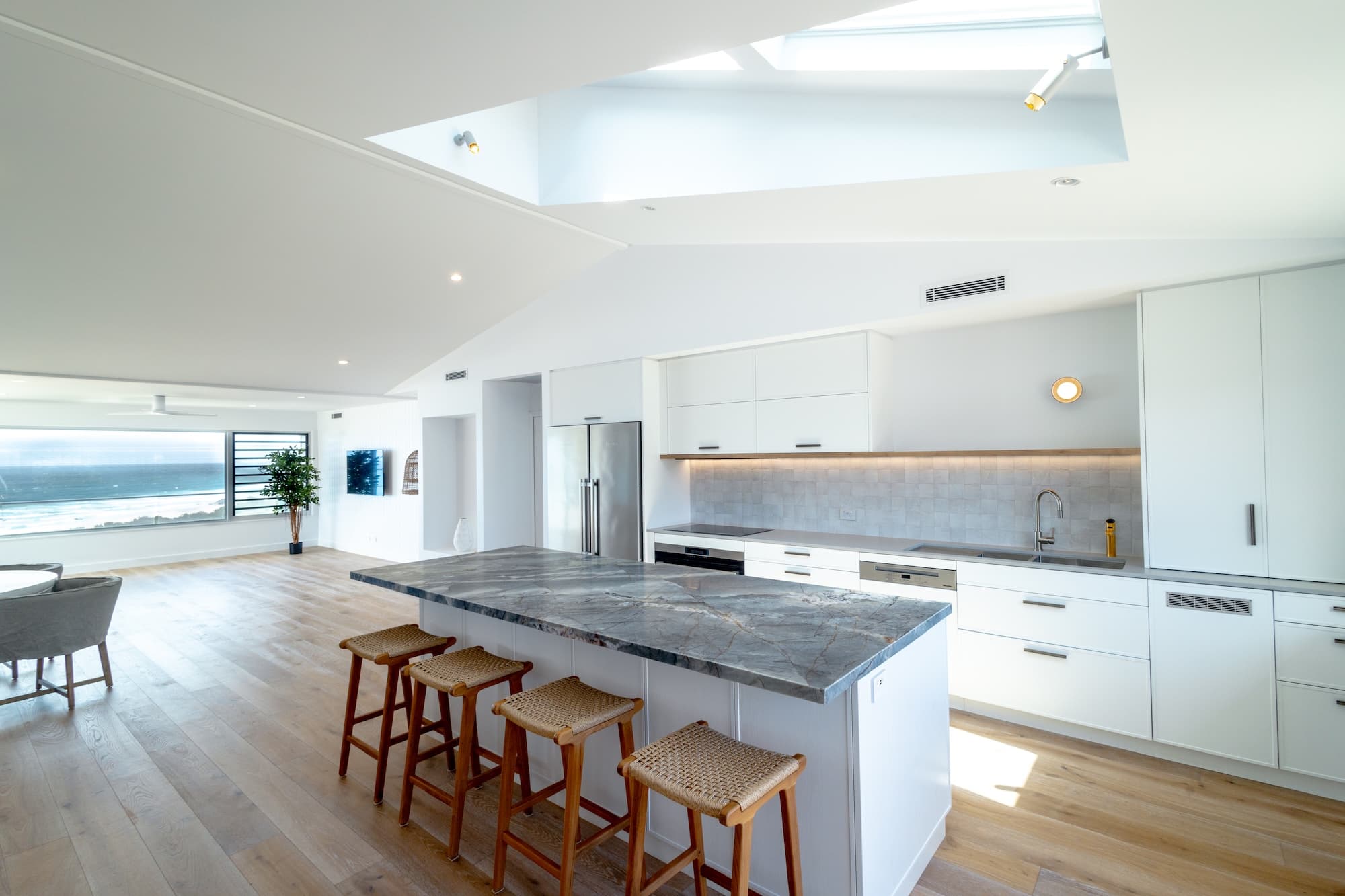 White coastal kitchen with a marble island, woven stools and an ocean view